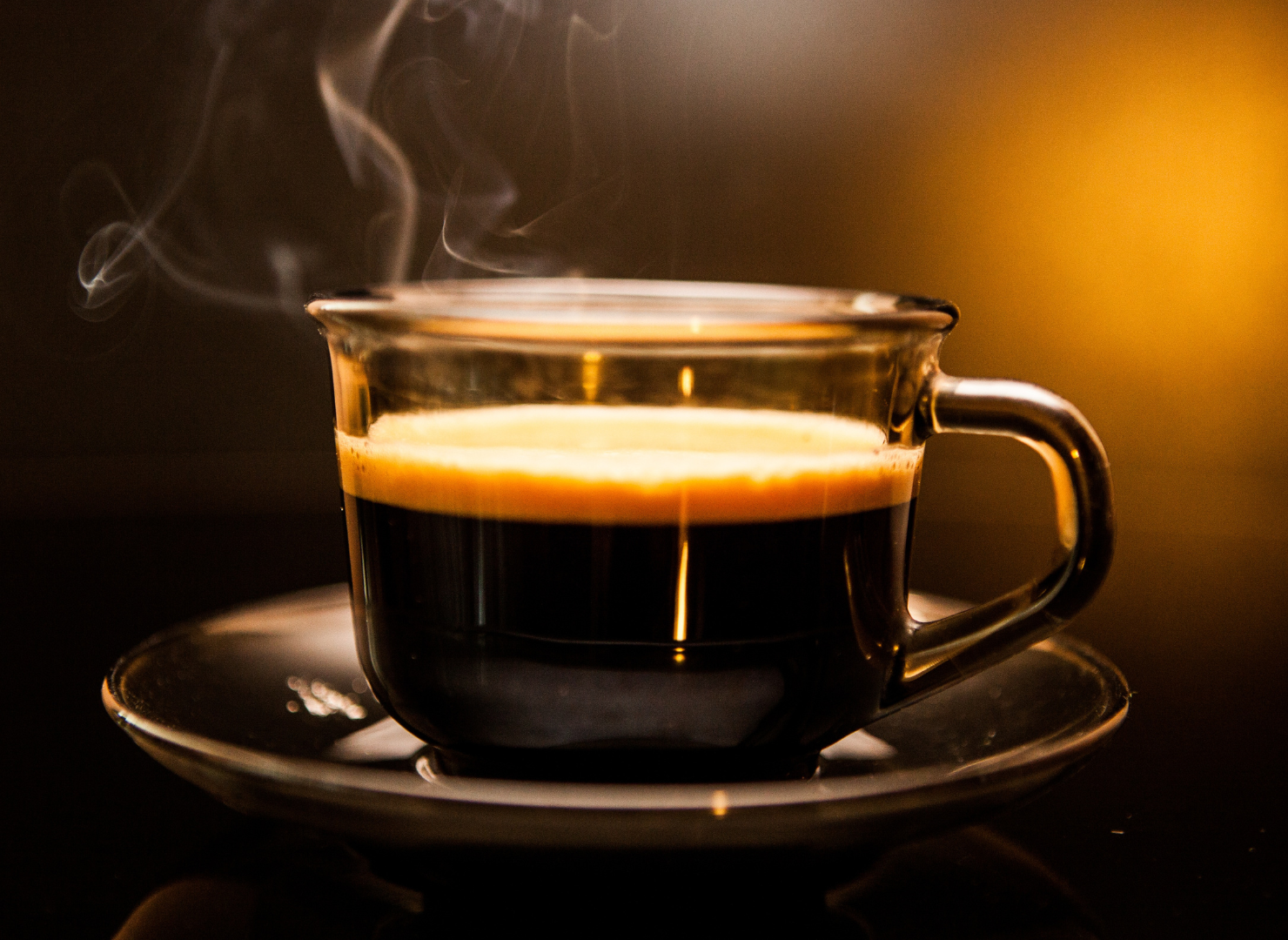 En glaskop med sort kaffe på en underkop med mørk baggrund.