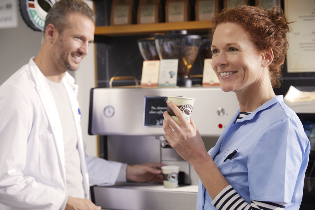 To personer ved en kaffemaskine – én i hvid kittel betjener maskinen, én i blå uniform holder en kaffekop.